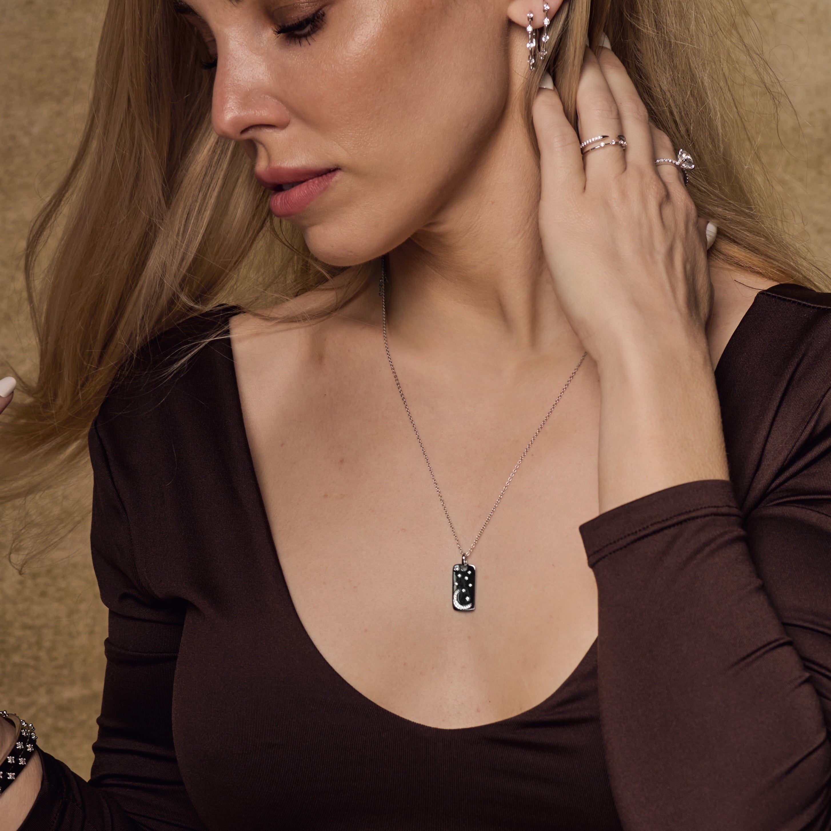 A woman in a deep brown, long-sleeved top touches her hair as she faces sideways, highlighting the Moonlight Pendant Necklace with a black accent, stacked rings, and silver earrings against a neutral background.