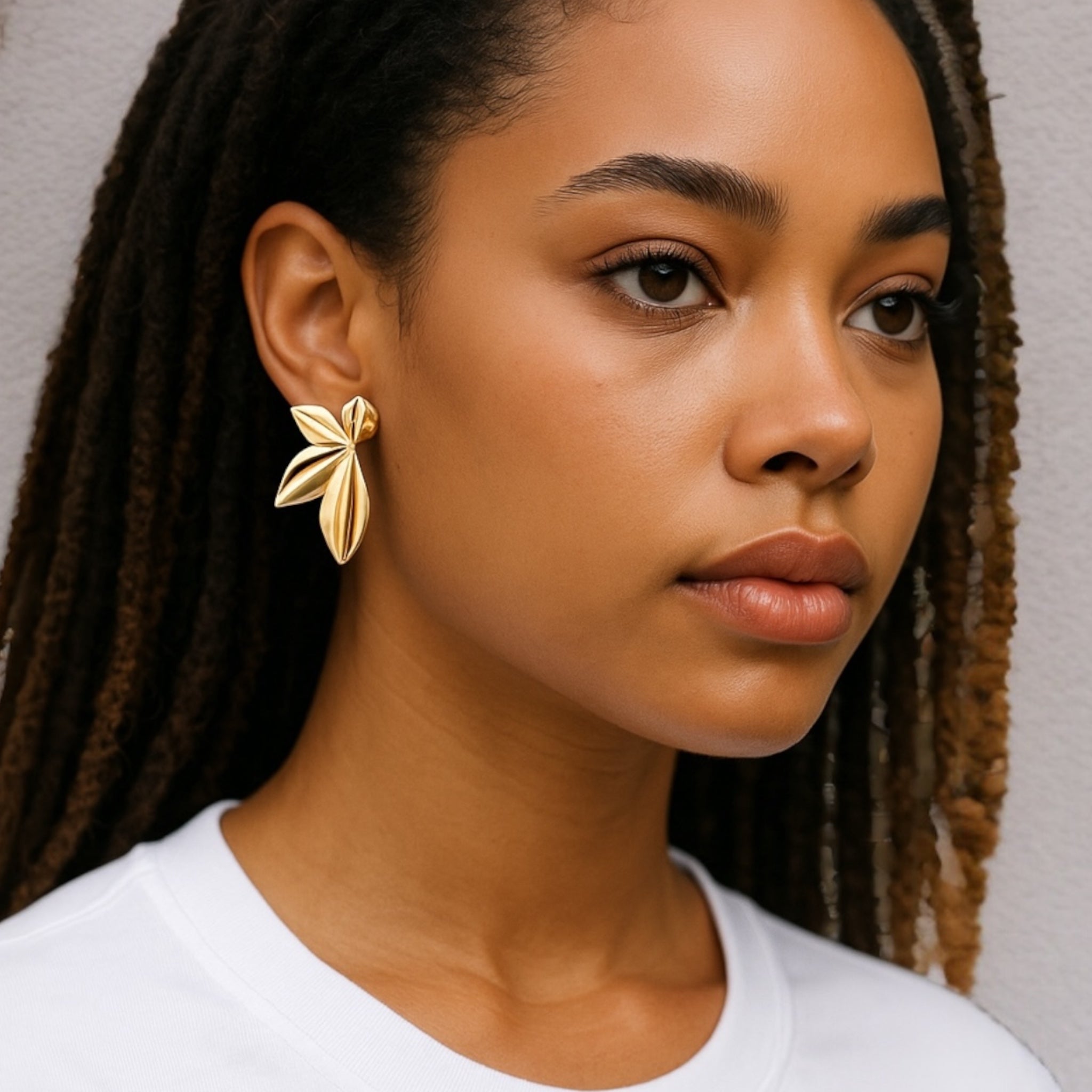 A woman with long, dark braids wears a white shirt and bold Harper Statement Petal Earrings in 14k gold plate. She stands against a neutral background, gazing calmly to the side.