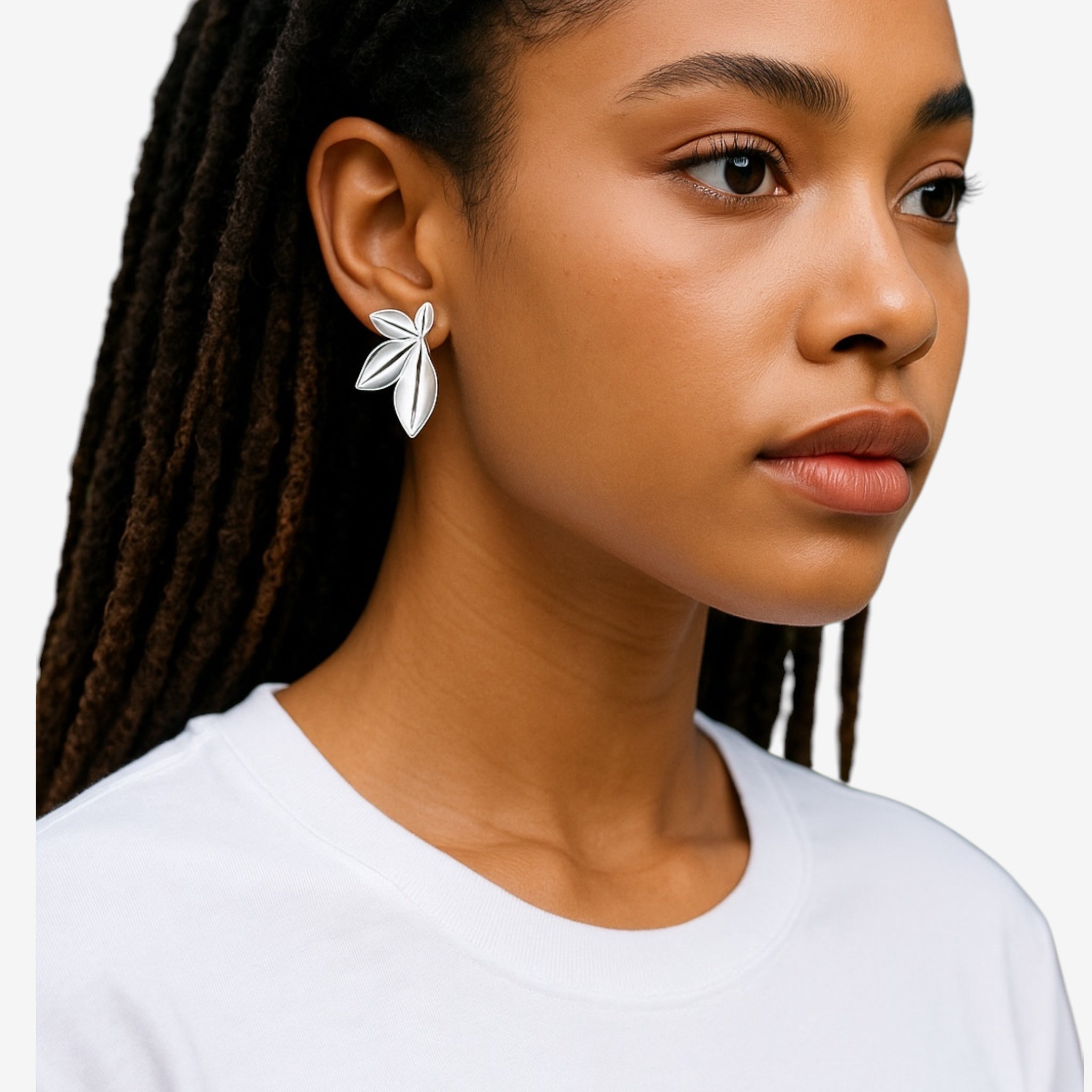 A woman with long, dark braided hair wears a white t-shirt and the Harper Statement Petal Earrings—oversized, silver leaf-shaped accessories—while gazing slightly to the side against a plain white background.