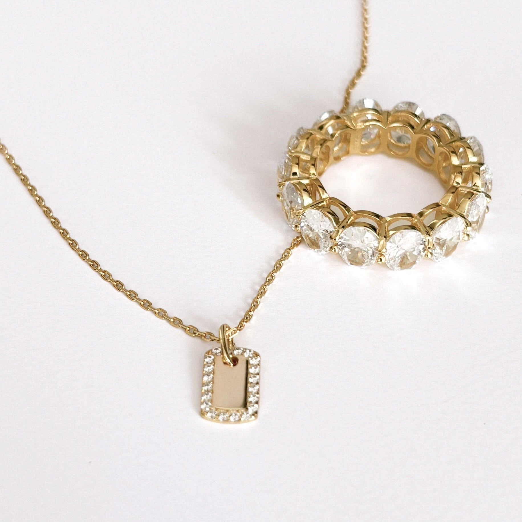 The Dainty Dog Tag Necklace – Gold is a 14k gold plated chain with a small rectangular pendant featuring tiny diamonds, shown beside a bold gold ring with large round-cut clear gemstones, both elegantly displayed on a white surface.