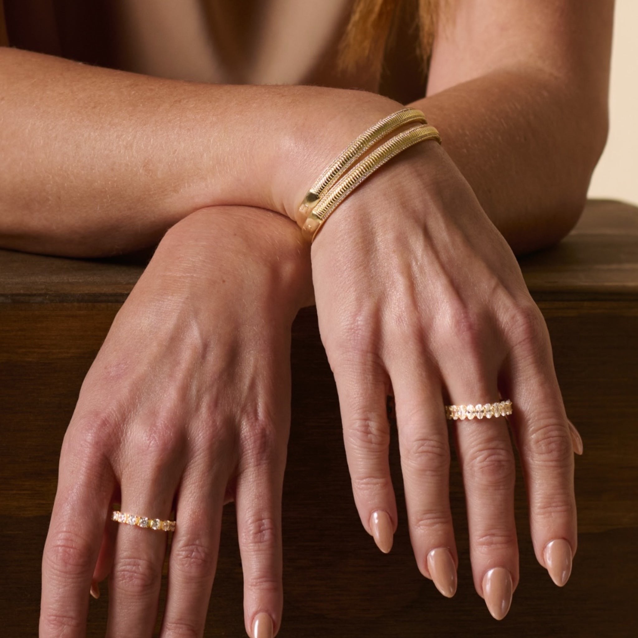 A womans manicured hands with nude polish rest on wood, showcasing the elegant Alyssa Fluted Bangle and gold rings with micro pave simulated diamonds.