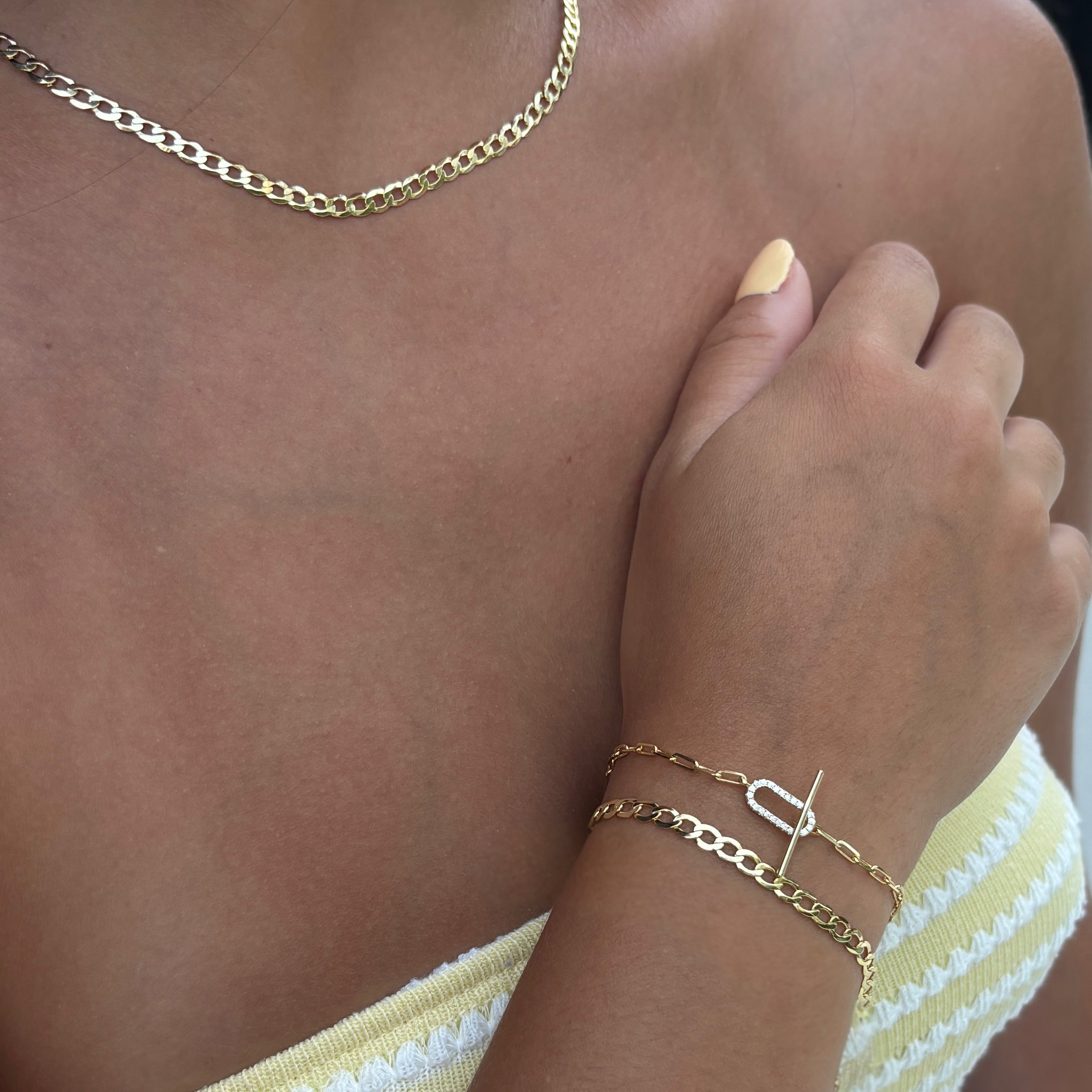 A person wearing a 14k gold plated chain necklace and two gold bracelets, including the Brynn Cuban Bracelet with a T-bar clasp, styled with a yellow and white striped top and pale yellow nail polish.