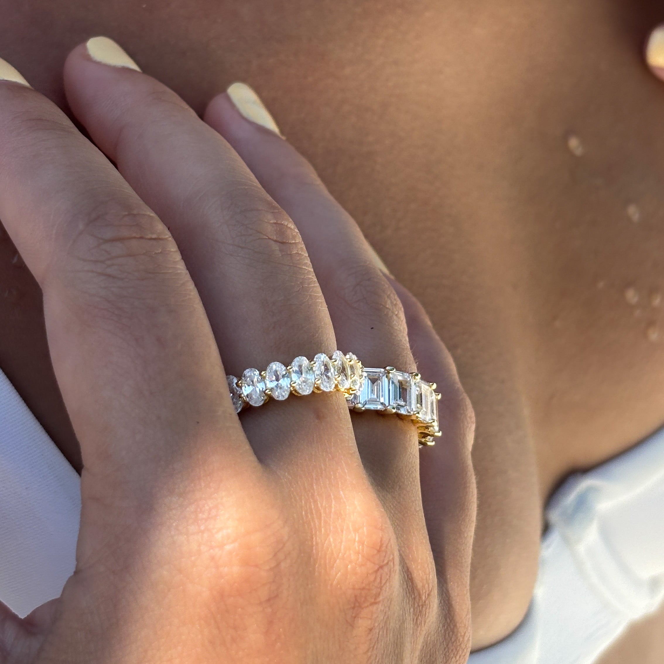 A close-up of a hand wearing two gold rings—one is the Aria Ring, adorned with oval-shaped stones—against sunlit skin and a white fabric background.