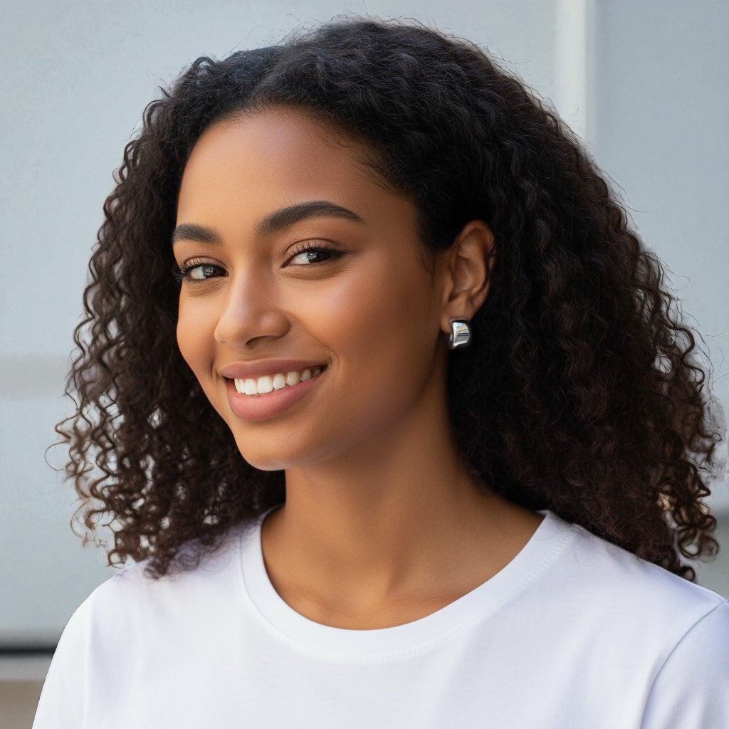 A young woman with curly hair smiles warmly, wearing a white shirt and the Brookes Curved Stud Earrings, standing in front of a light-colored background.