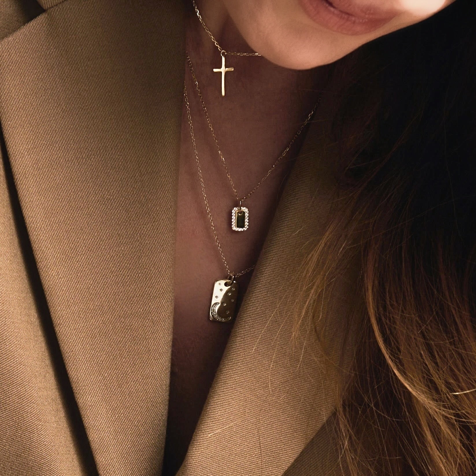 A close-up of a woman in a brown blazer wearing three layered gold necklaces, including the Dainty Dog Tag Necklace, with her hair partially visible on the right.