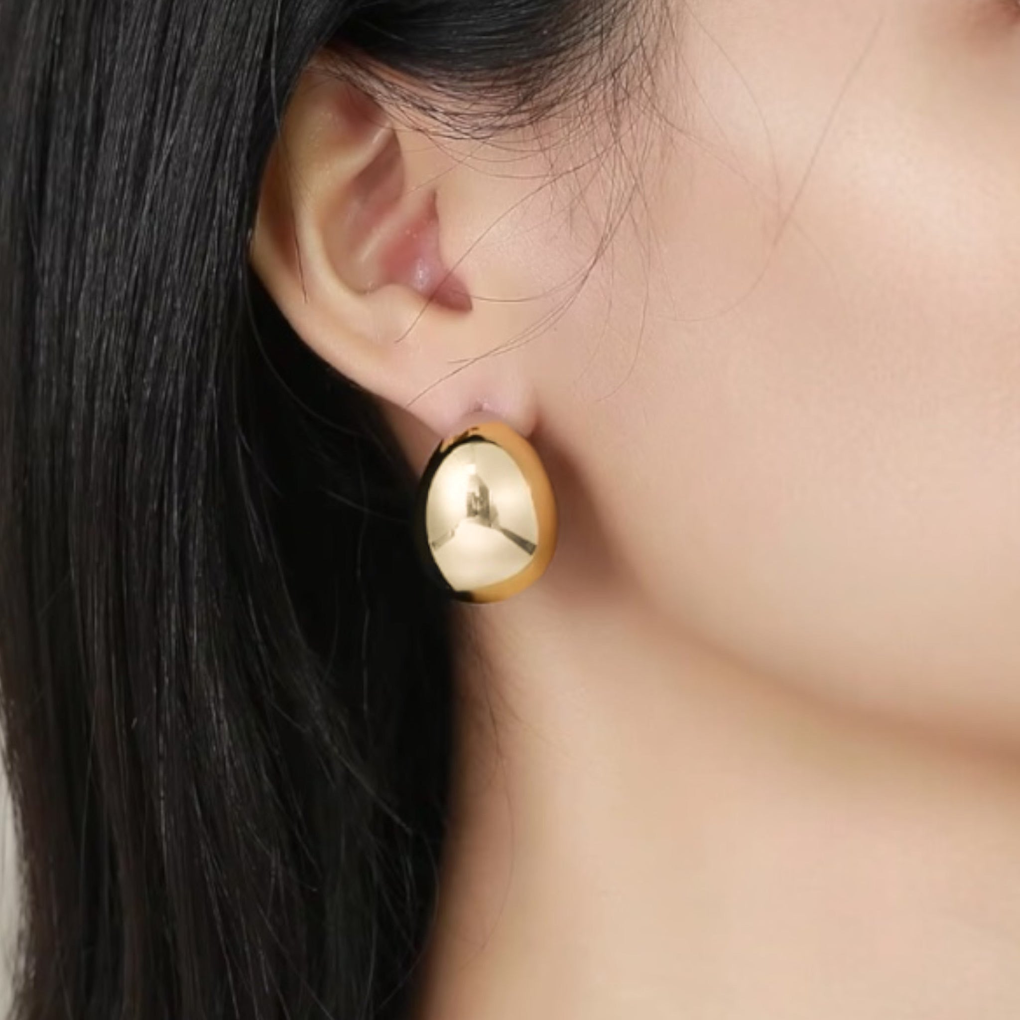 Close-up of a person with straight black hair and smooth, light skin wearing shiny Dani Crescent Earrings. The oval-shaped, 14k gold plated statement earrings stand out against the softly blurred neutral background.