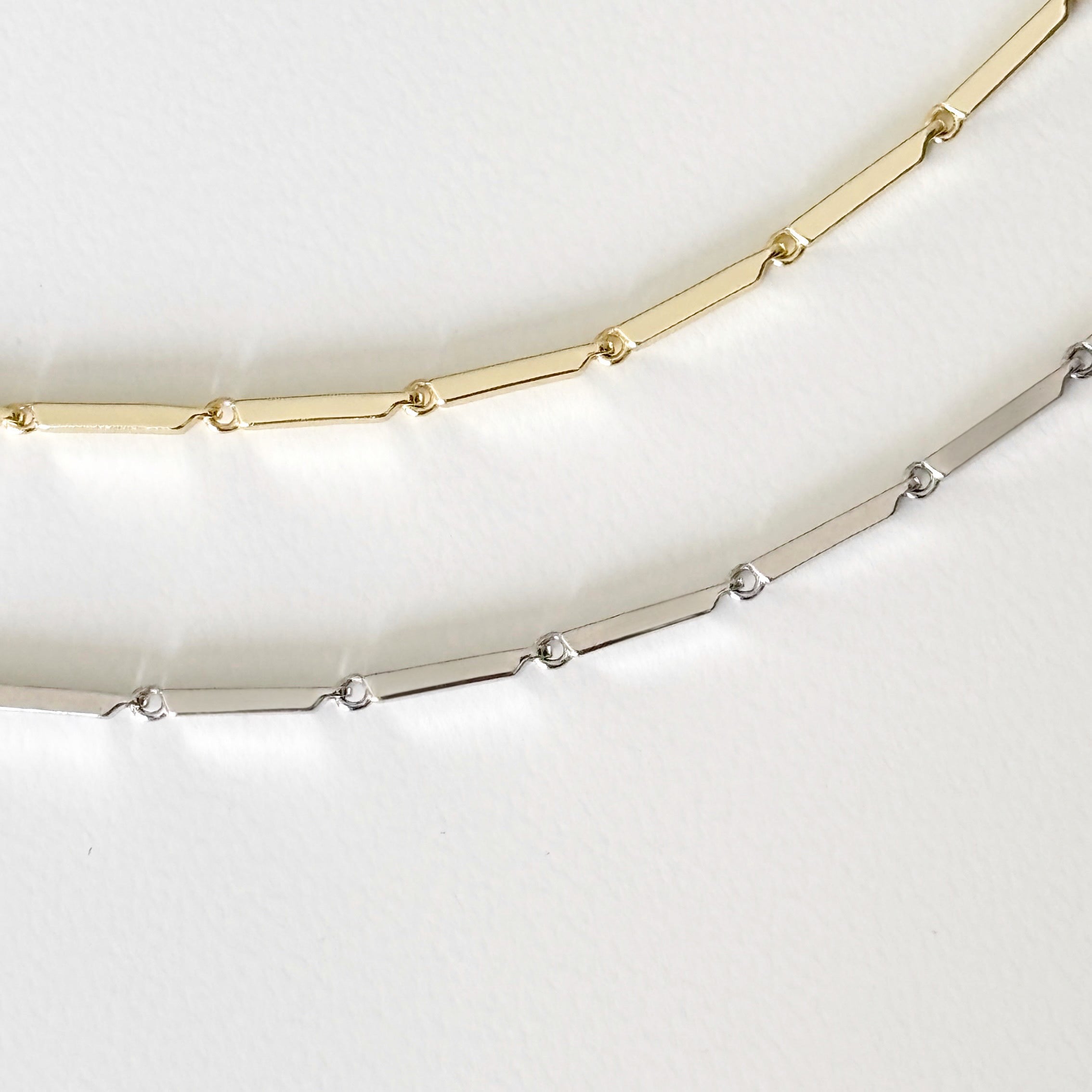 Two Emmie Necklaces rest on a white surface: one in gold, the other in sterling silver. Both feature elongated rectangular links joined by small round links, creating an elegant and modern minimalist look.