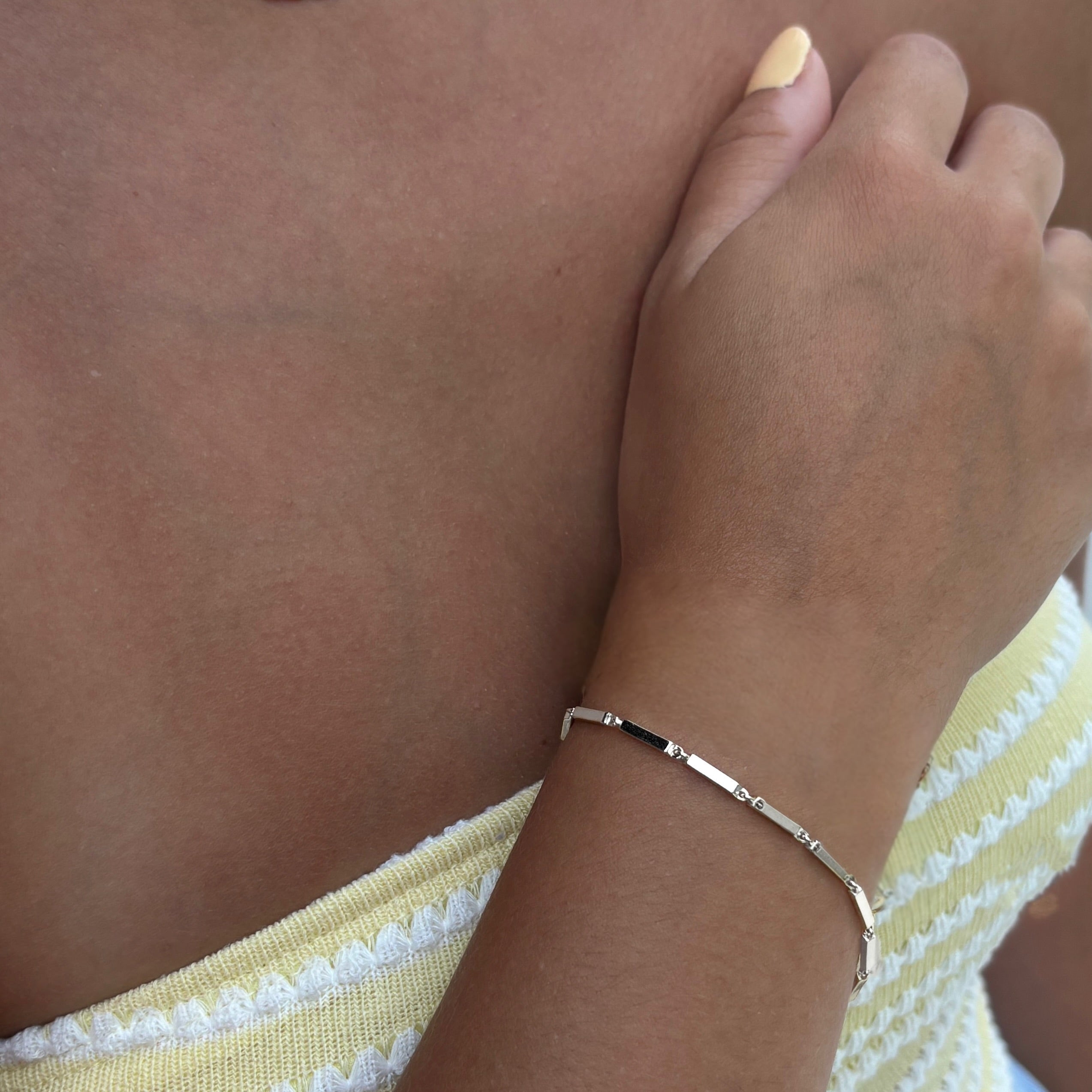 A person with light skin wears a yellow and white textured strapless top and the delicate Emmie Bracelet, holding one hand near their shoulder. Their nails are painted a soft yellow.