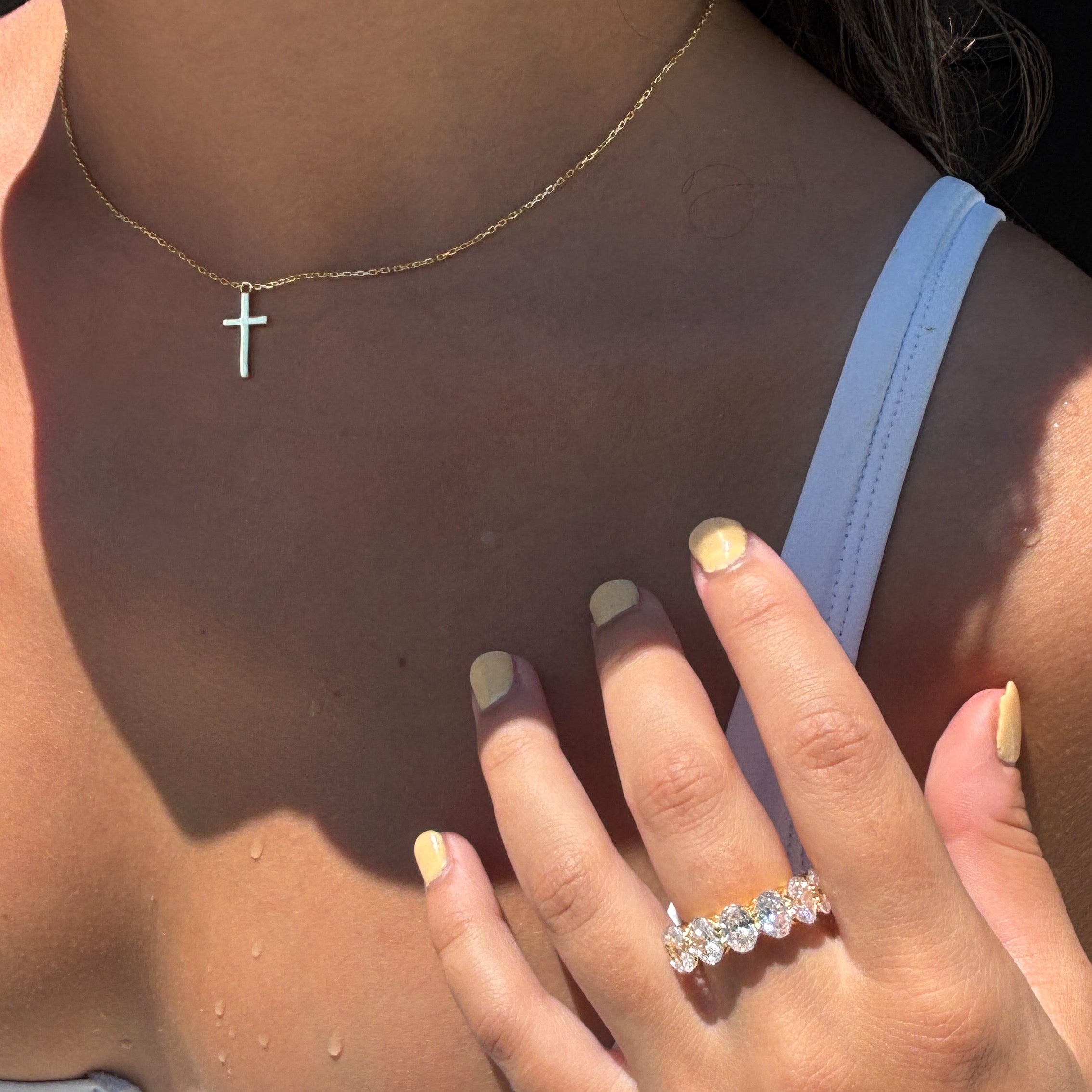 A person wears the Classic Cross Necklace with a white top, light yellow nail polish, and a ring with five clear stones. Water droplets are visible on their shoulder and chest.