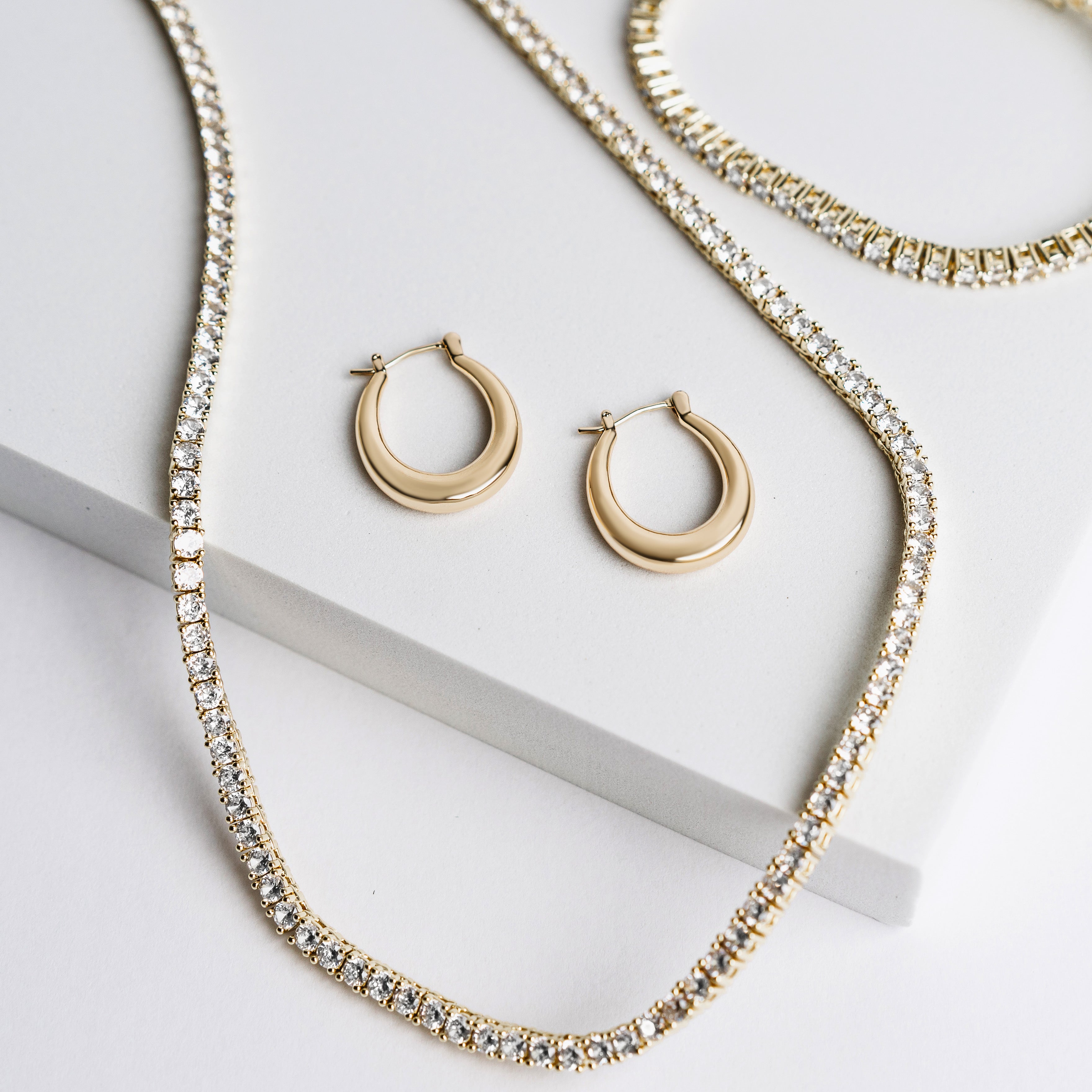 Riley Oval Hoops and two sparkling gold necklaces with clear gemstones are displayed on white geometric surfaces in a minimal, elegant setting—perfect for everyday wear.