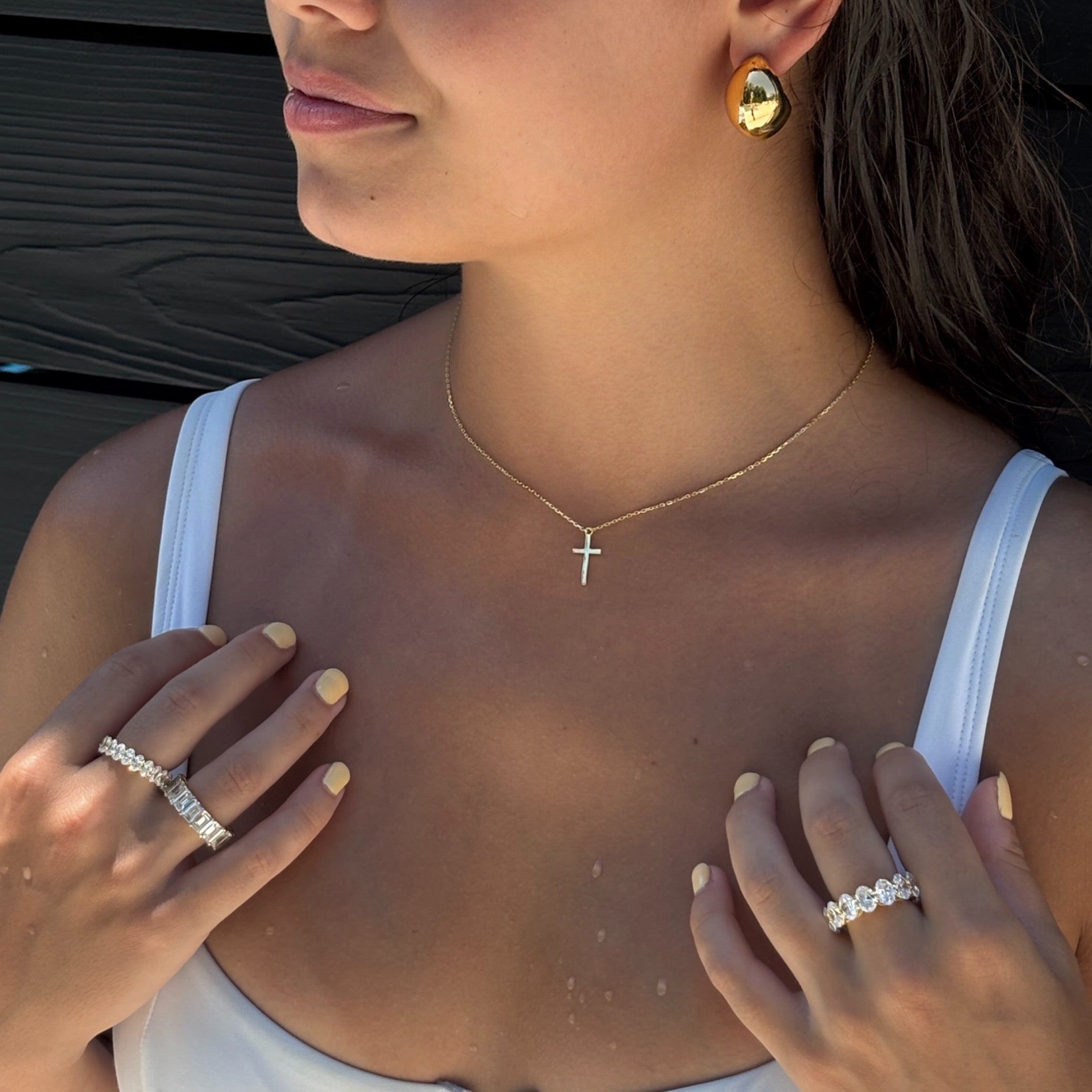 A woman in a white top showcases Dani Crescent Earrings, a delicate gold cross necklace, sparkling diamond rings on each hand, and light yellow nail polish.