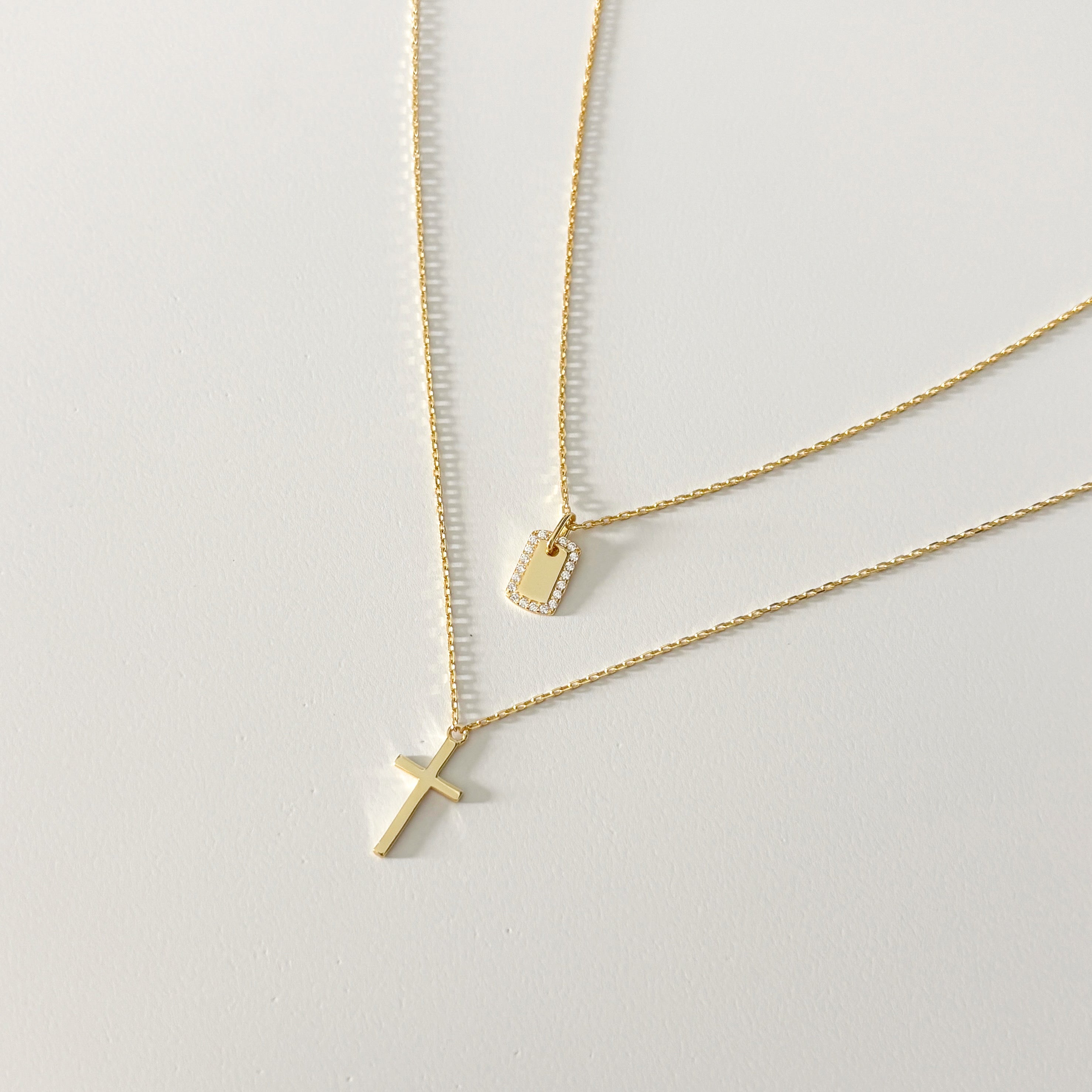 Two delicate gold necklaces on a white background—one features a 14k gold pendant, the other is the Classic Cross Necklace. Both chains are thin and minimalist, perfect for adding everyday elegance to your look.