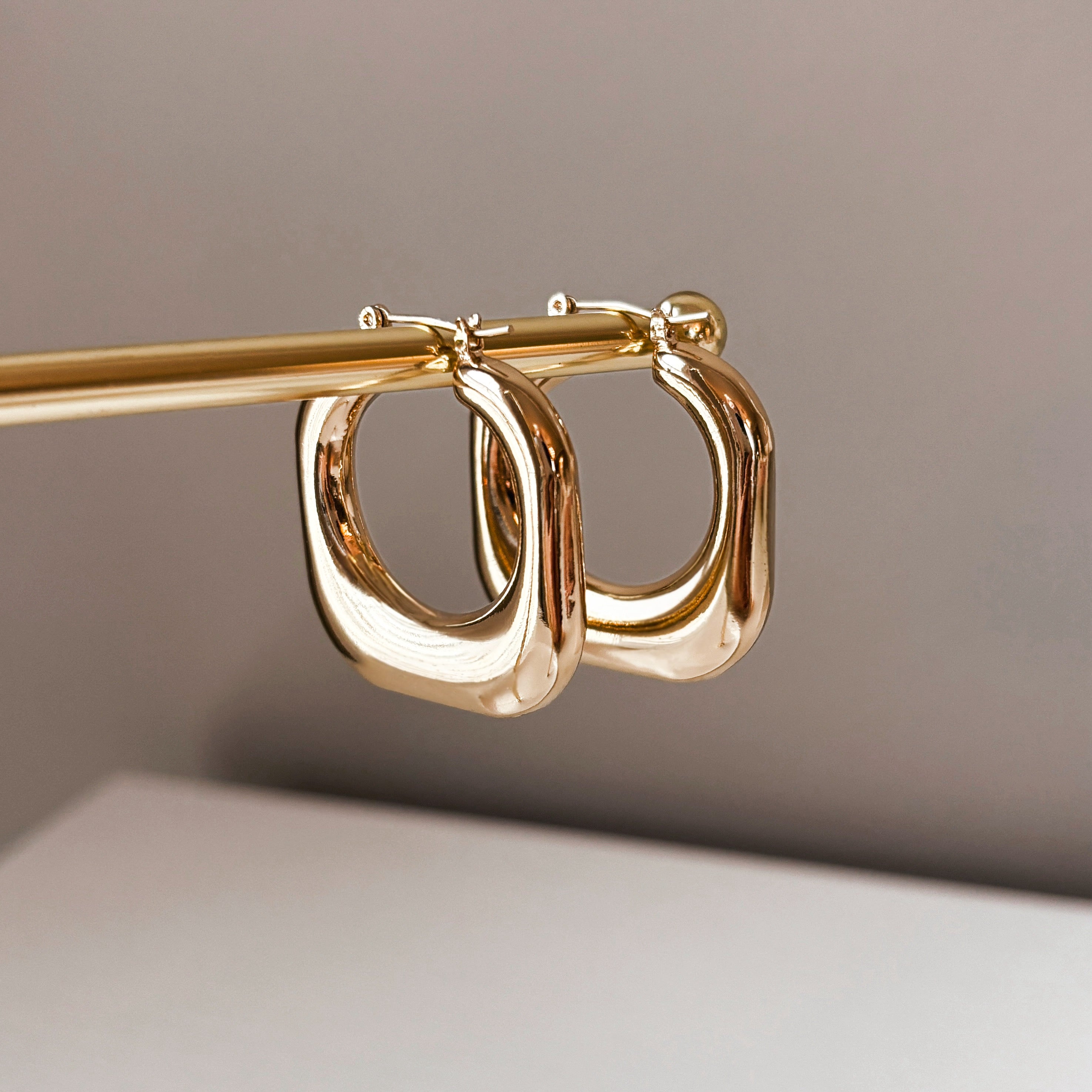 The Stormy Squared Hoops, a pair of shiny modern gold earrings with a square shape, hang from a gold rod against a neutral background.