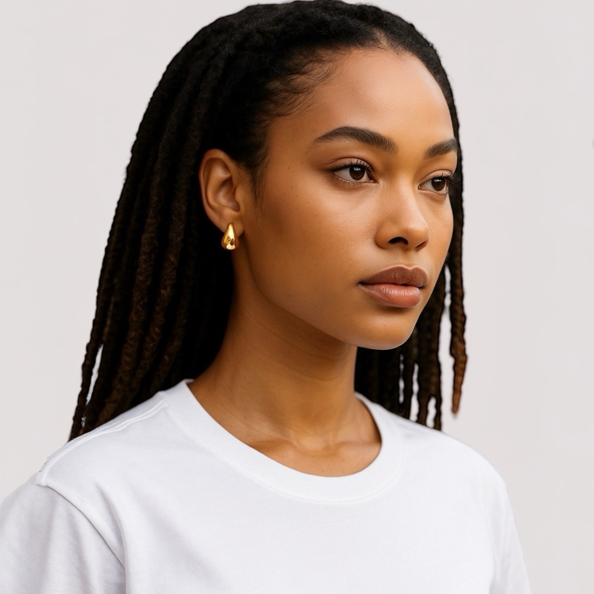 A woman with long, dark, braided hair wears Demi Teardrop Earrings and a plain white t-shirt as she gazes thoughtfully into the distance against a neutral background.