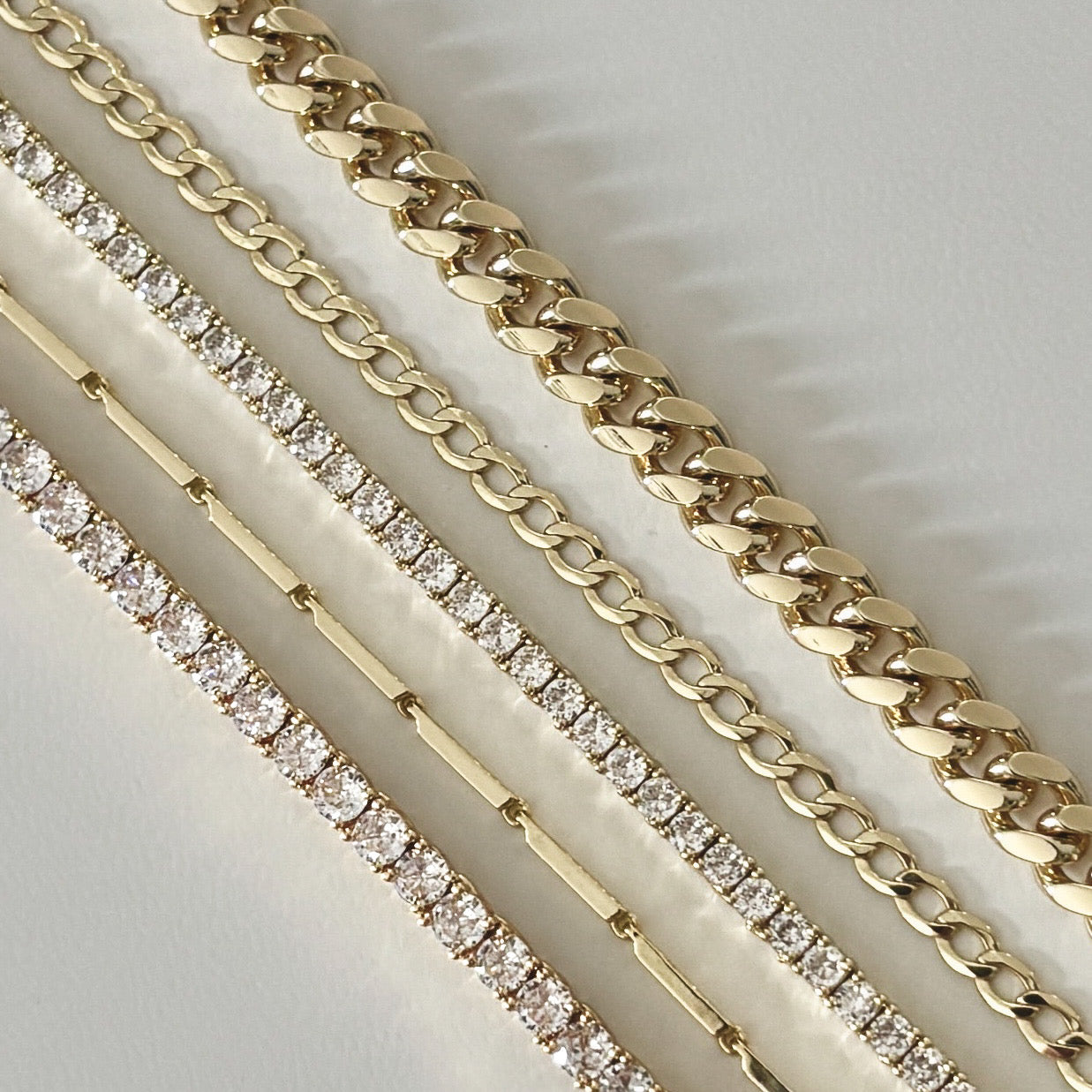 Four gold chain bracelets rest diagonally on a white surface; two showcase plain links, while two shimmer with round-cut diamonds—perfectly paired with the Emmie Necklace for an elegant, layered gold look.