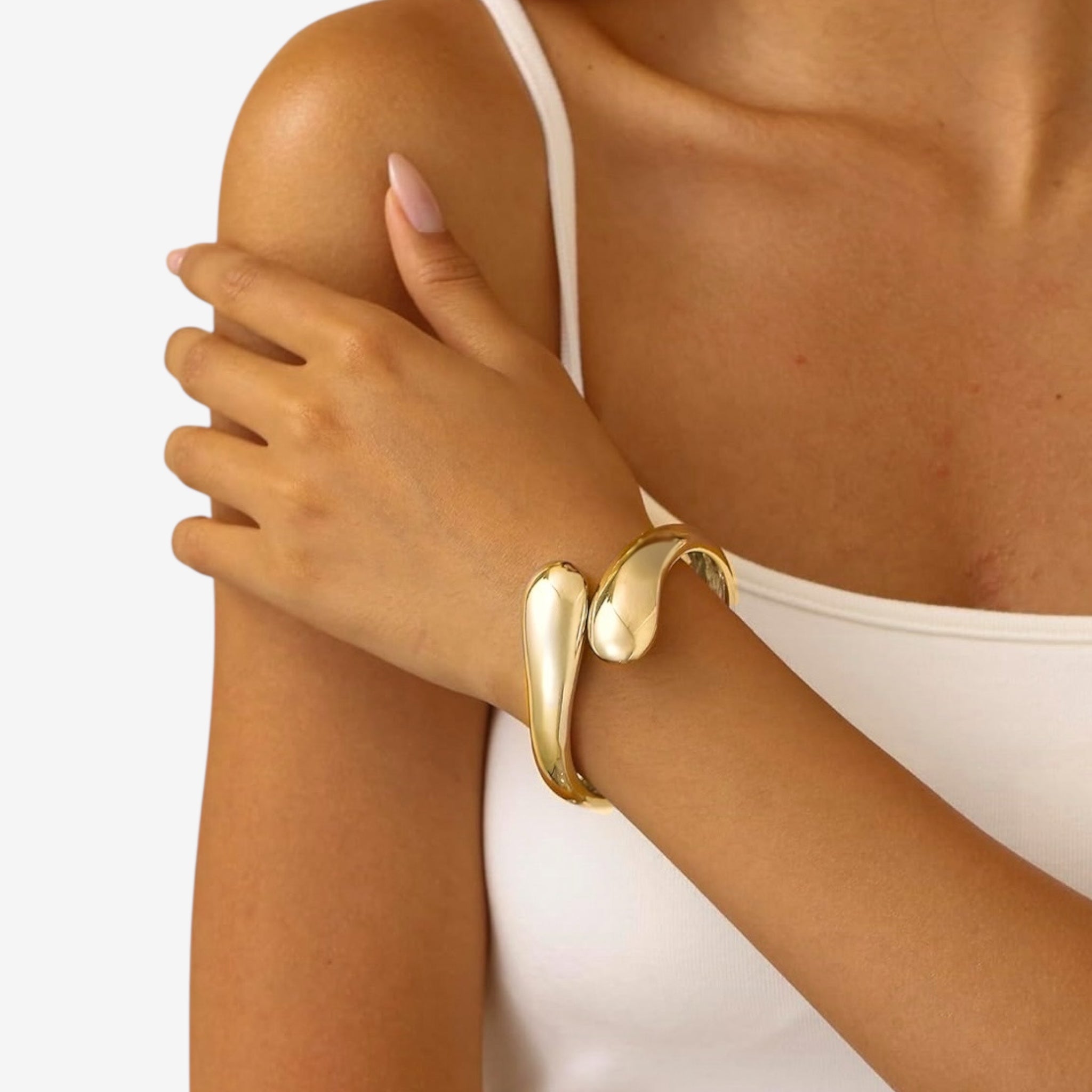 A woman in a white spaghetti strap top rests her hand on her shoulder, highlighting the Jade Snake Bangle—a shiny, sculptural gold statement bracelet. Her nails are manicured in a nude shade.