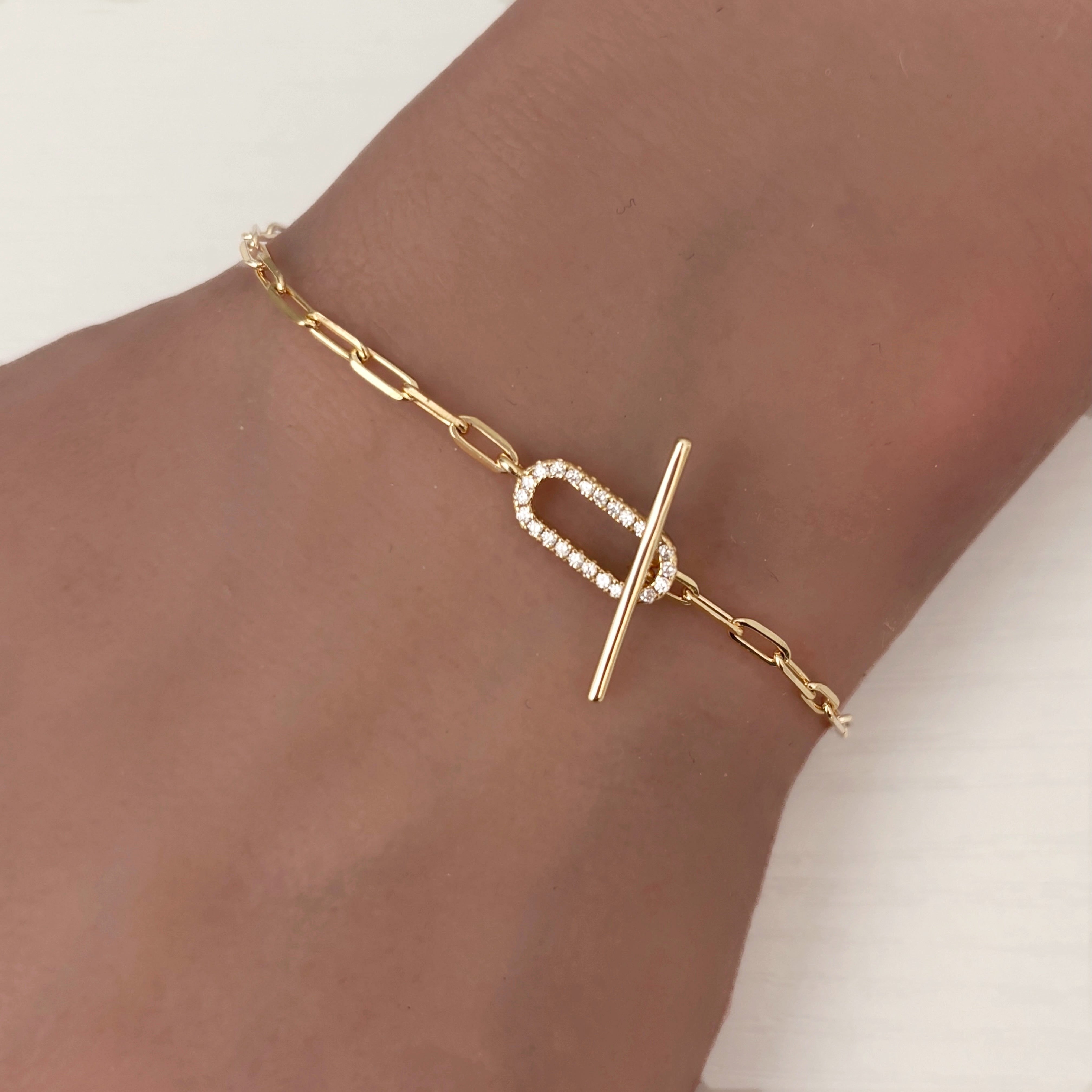 The Jules Bracelet is a gold paperclip chain bracelet with a toggle clasp and a U-shaped section accented by sparkling clear stones, shown worn on a wrist against a light background—an elegant accessory.