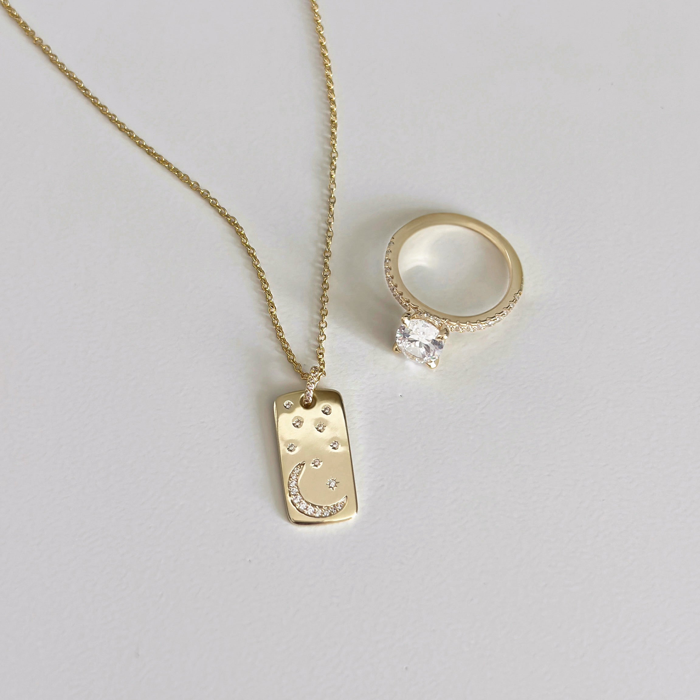 The Moonlight Pendant Necklace, featuring a rectangular gold pendant with a crescent moon and star design, is displayed beside a gold ring with a large round clear gemstone on a light background.