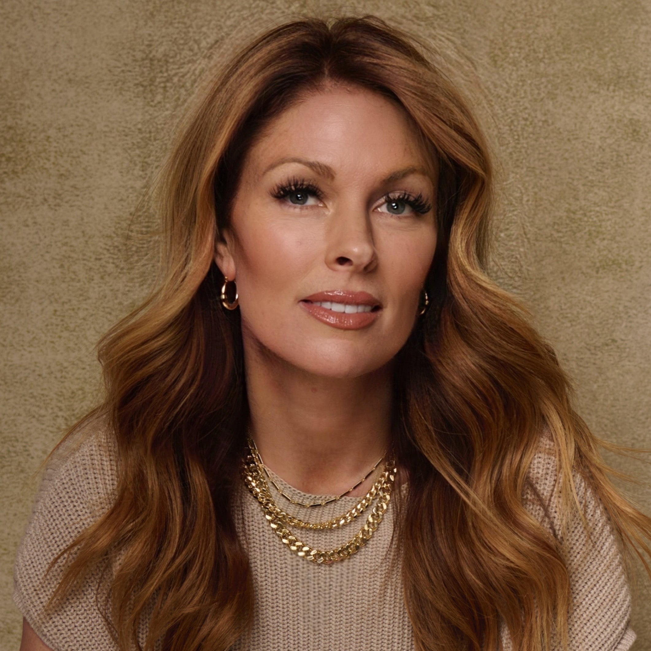A woman with long, wavy, light brown hair in a beige top smiles softly, her Riley Oval Hoops and layered necklaces glowing against the textured beige background.