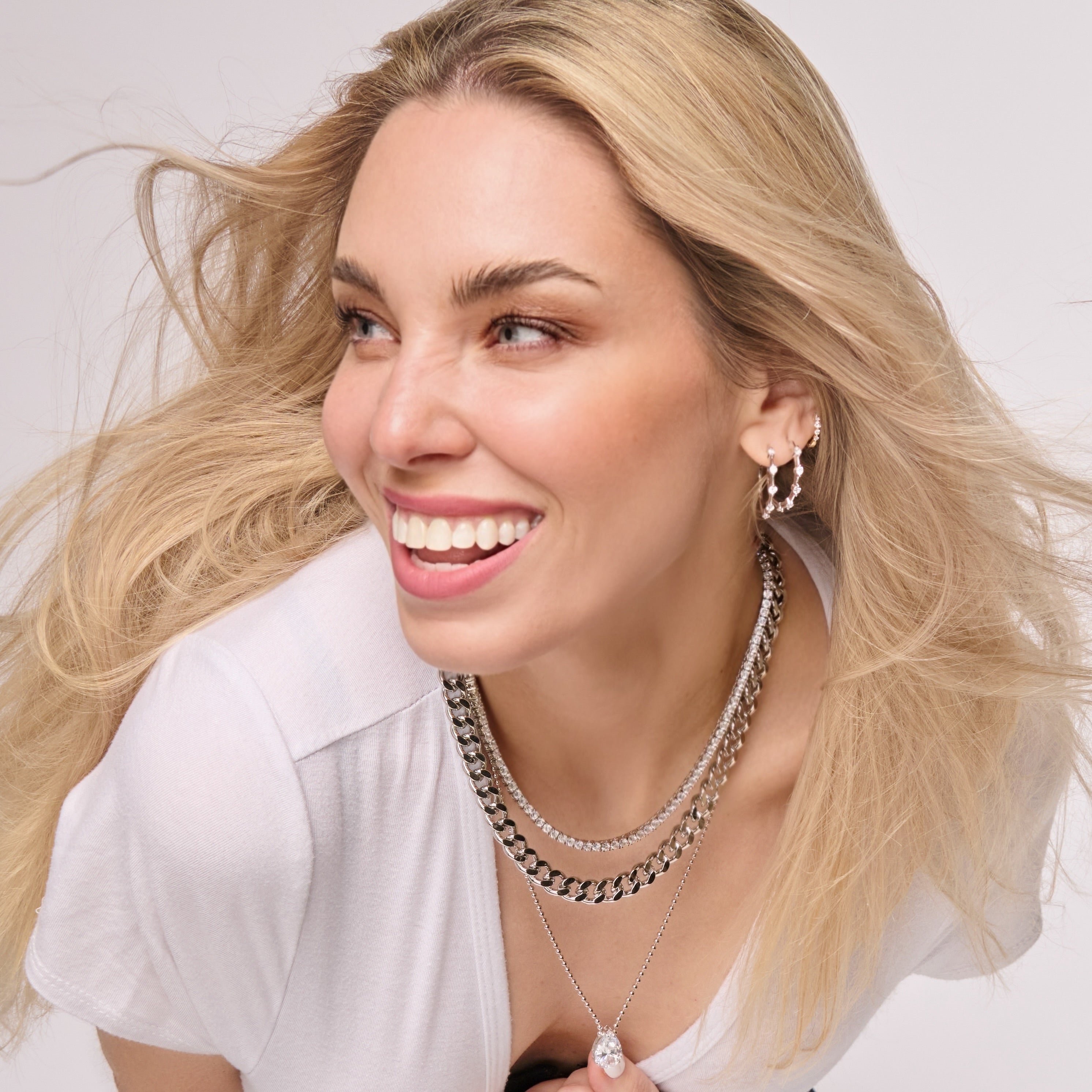 A smiling woman with long blonde hair wears the Zoe Statement Curb Chain layered with silver pieces, hoop earrings, and a white top, looking slightly to the side against a light background.