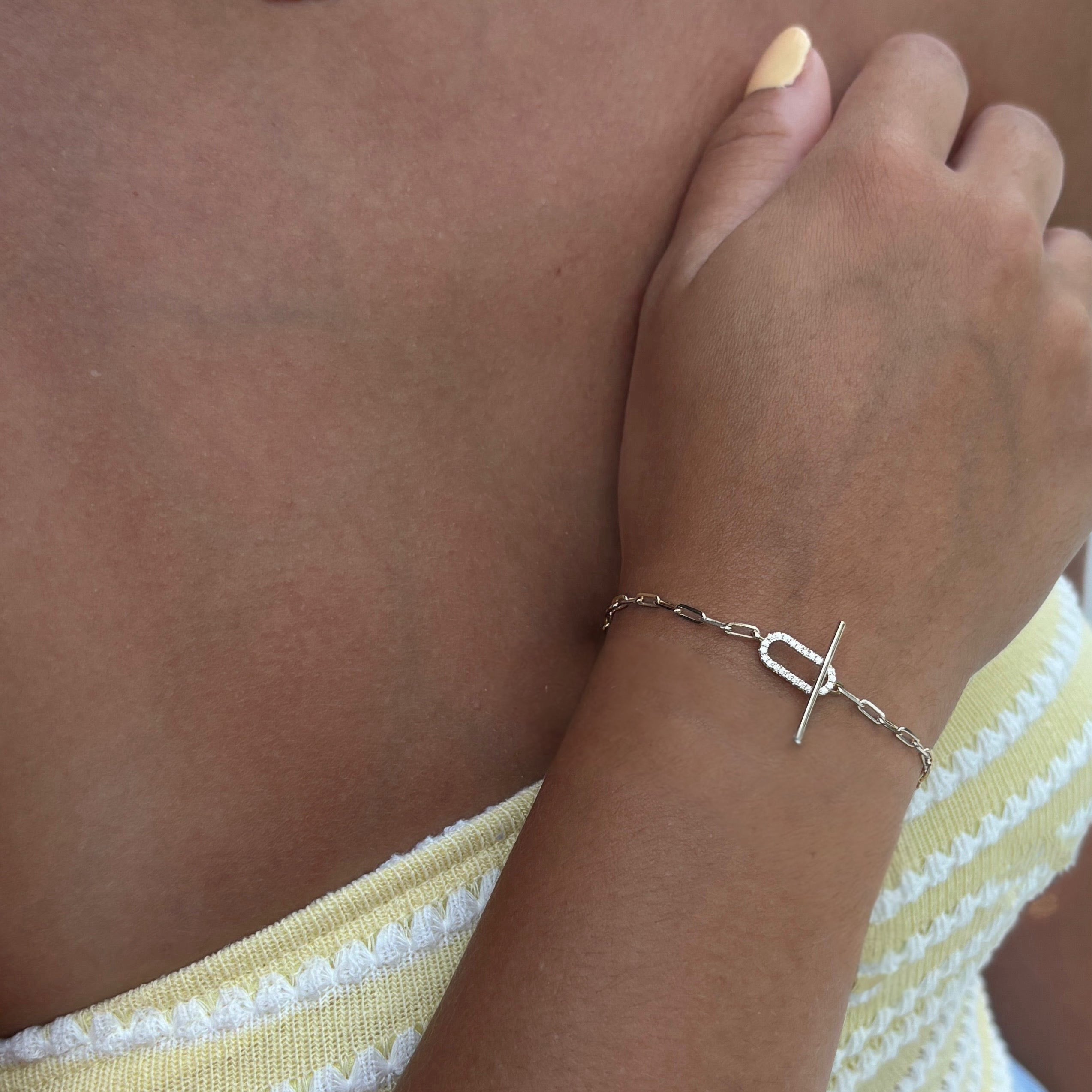 A close-up of model wearing the Jules Bracelet, a gold paperclip chain with a T-bar toggle clasp, on their wrist. They are dressed in a yellow and white striped top and have pale yellow painted nails.
