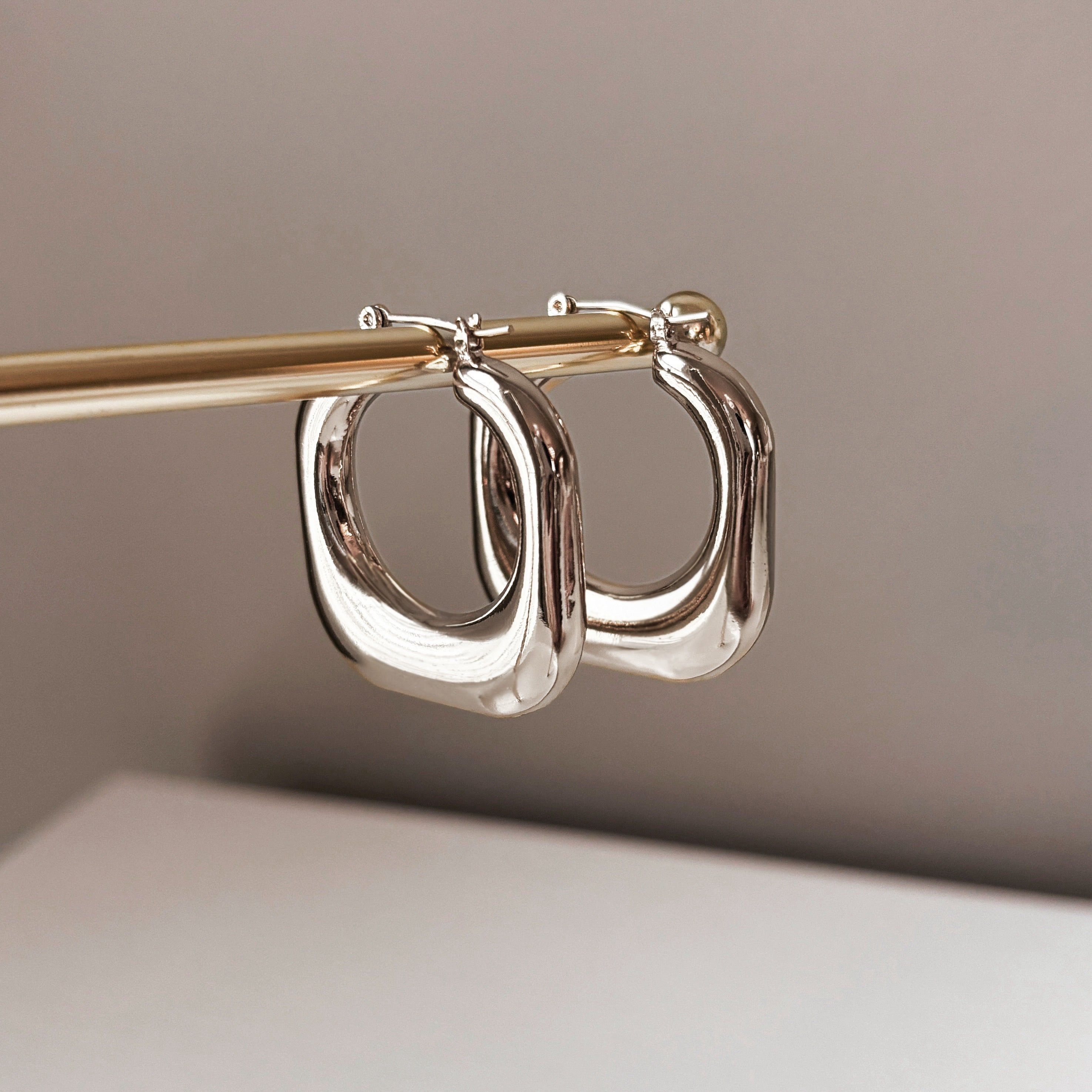 The Stormy Squared Hoops, bold silver earrings, are displayed hanging from a gold rod against a simple neutral background.