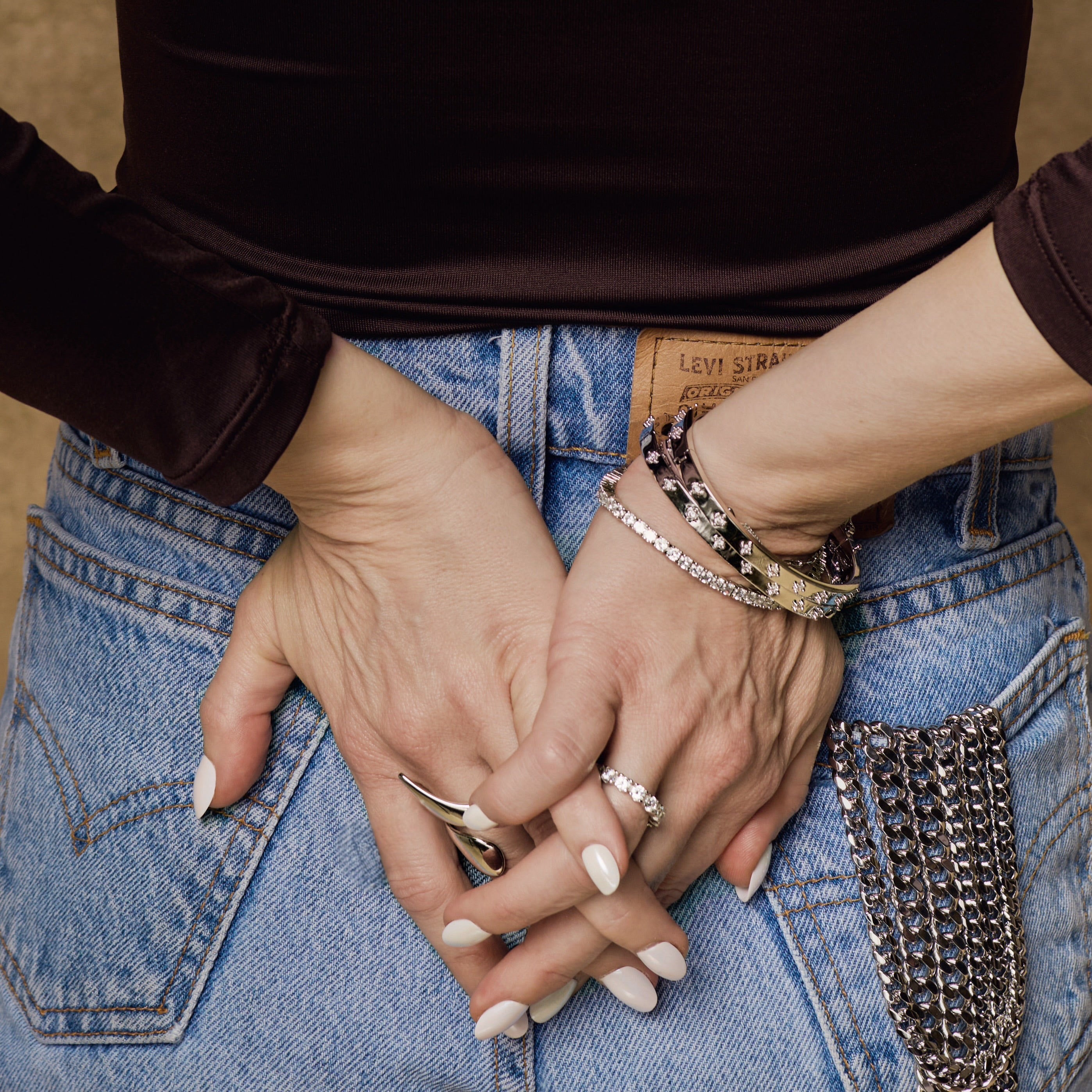 A person in a black long-sleeve top and blue Levi’s jeans with a chain detail stands with hands clasped behind their back, showcasing the Sydney Ring and several bracelets.