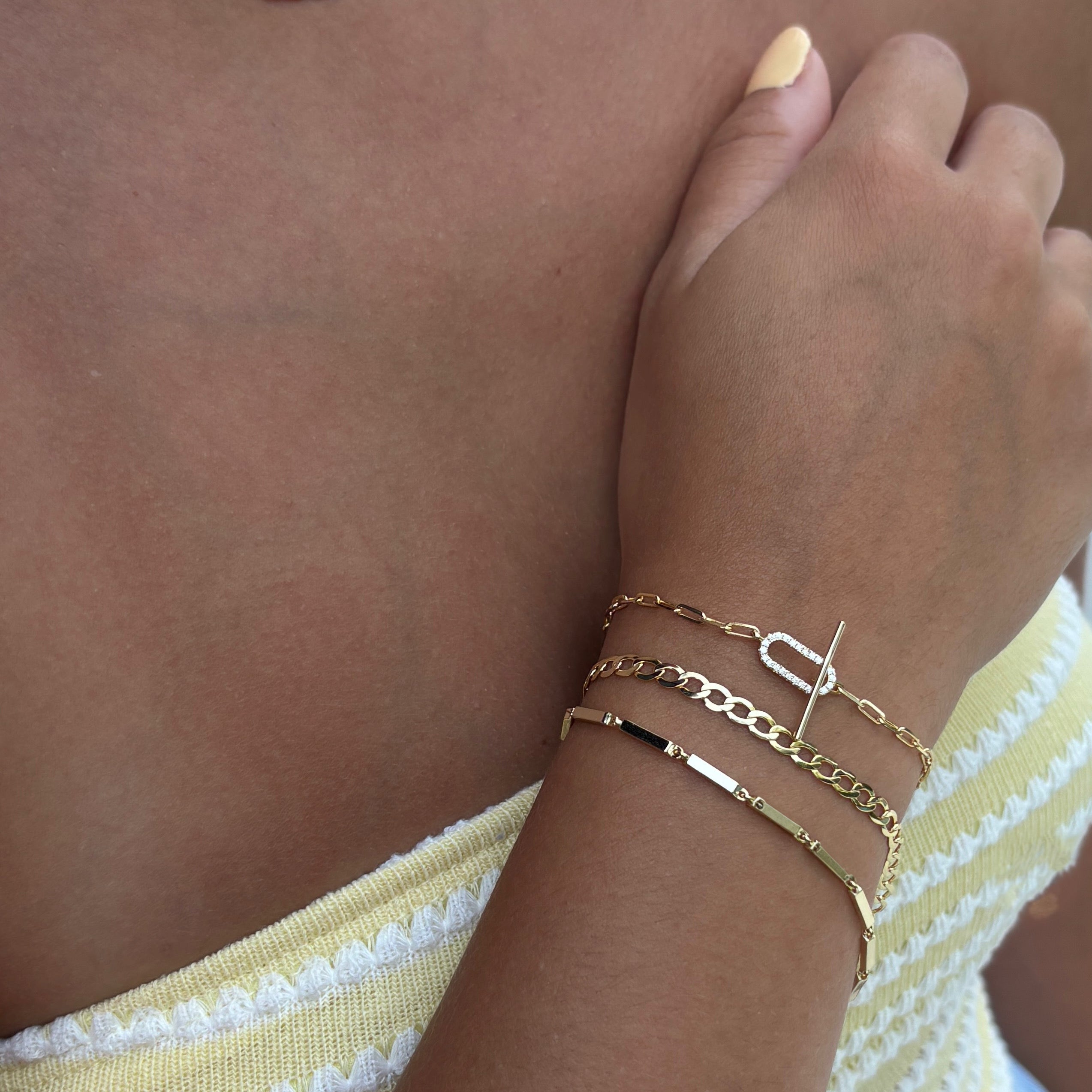 A person with light brown skin and pale yellow manicured nails wears the Jules Bracelet, a gold paperclip chain with a simulated diamond clasp. They’re dressed in a yellow and white textured strapless top; focus is on their arm and jewelry.