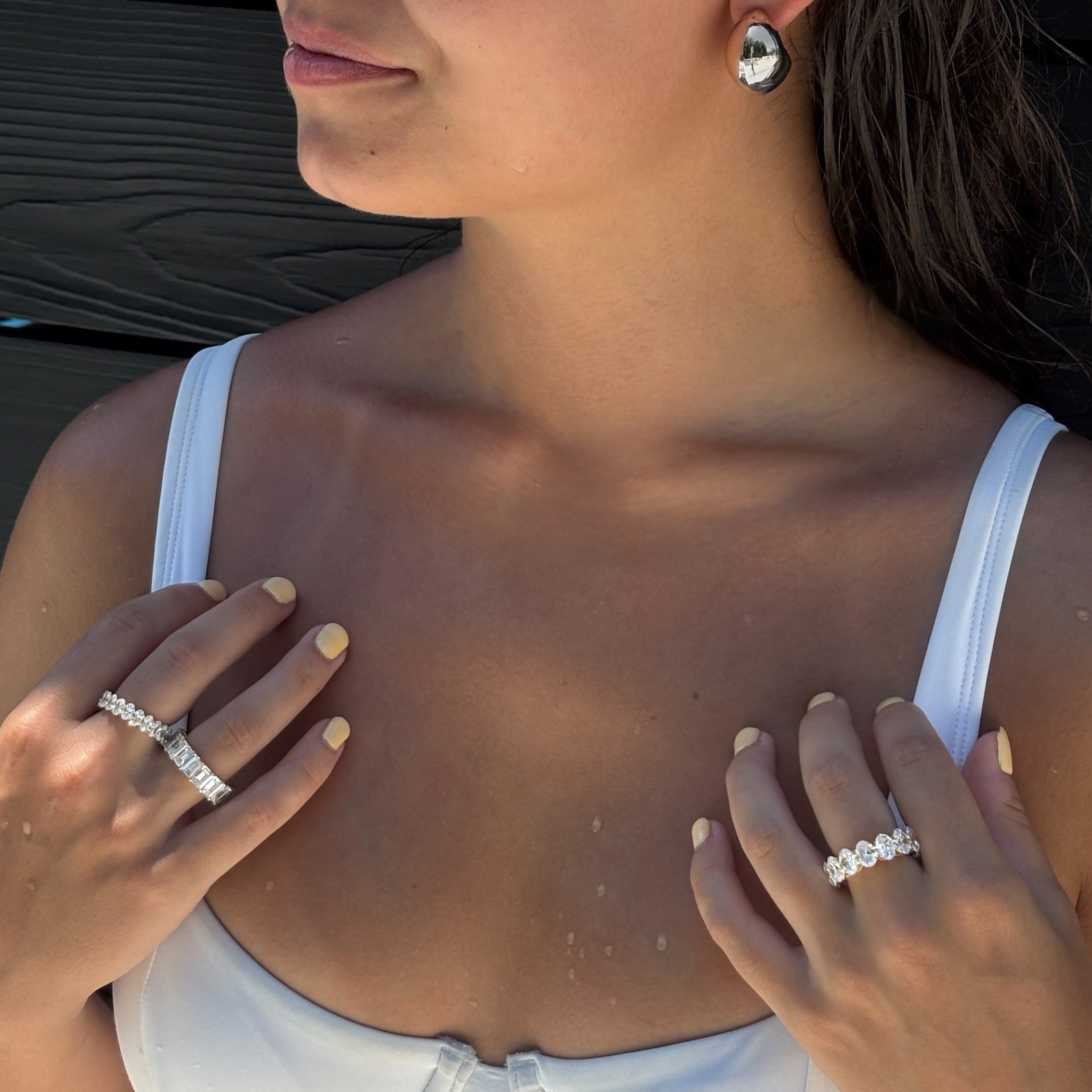 A woman wearing a white top showcases two Bella Rings and 925 sterling silver earrings with simulated diamonds. Her light yellow nails stand out as she poses with her hands near her collarbone against a dark backdrop.