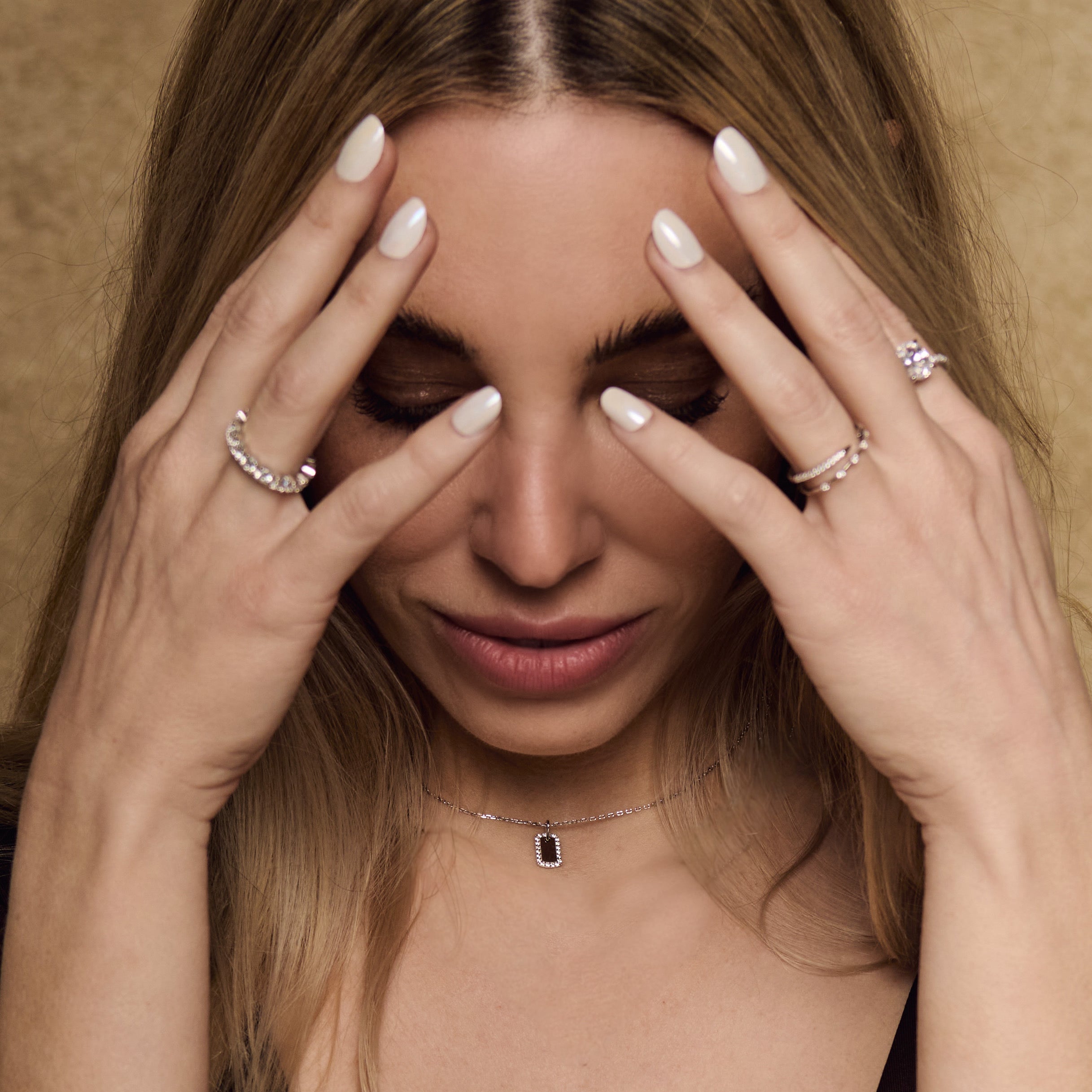 A woman with long blonde hair touches her forehead with both hands, eyes closed. She wears white nail polish, rings, and a sterling silver Dainty Dog Tag Necklace featuring a sleek rectangular pendant.