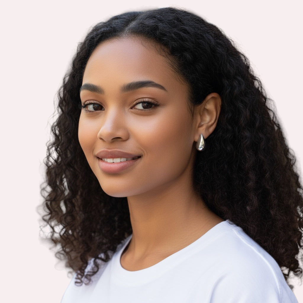 A young woman with curly dark hair smiles softly in a white shirt and Demi Teardrop Earrings, posed before a neutral background—a simple, stylish look that showcases the Demi Teardrop Earrings as perfect everyday jewelry.