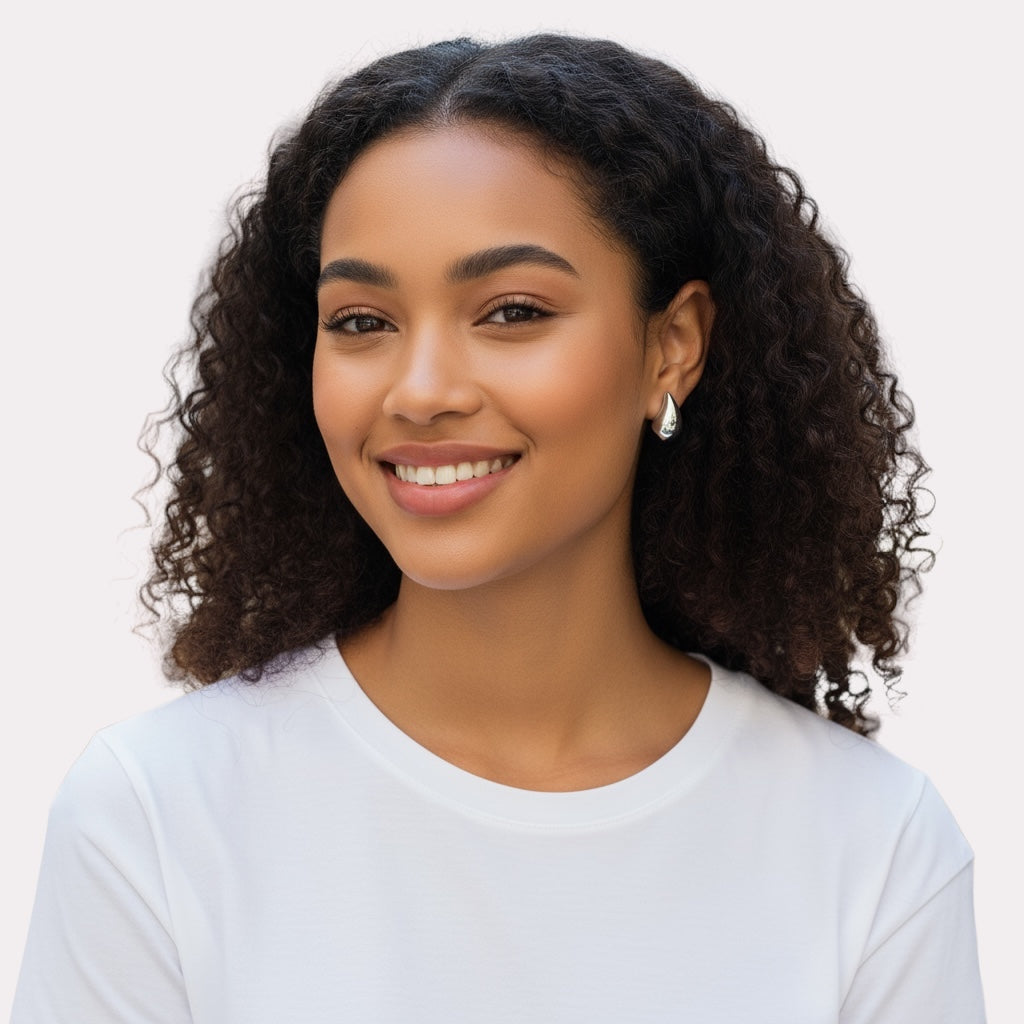 A young woman with curly dark hair and a white shirt smiles warmly against a plain light background, elegantly showcasing Demi Teardrop Earrings as her stylish choice for everyday jewelry.