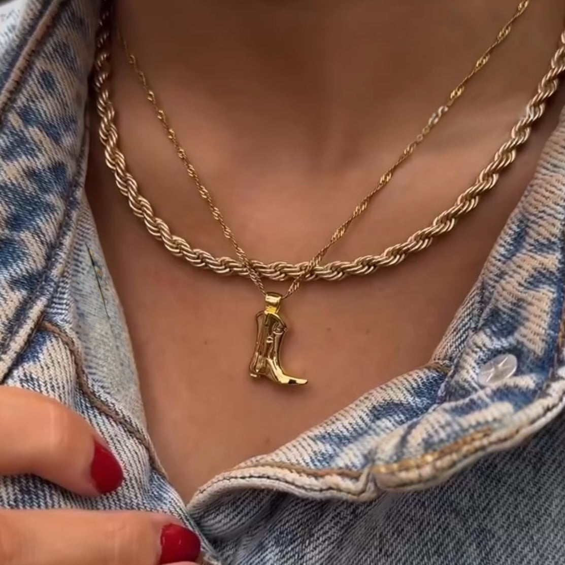 A close-up of a person wearing two gold necklaces—a twisted rope chain and the Boots & Broadway Pendant Necklace—styled with a denim jacket and red nail polish.