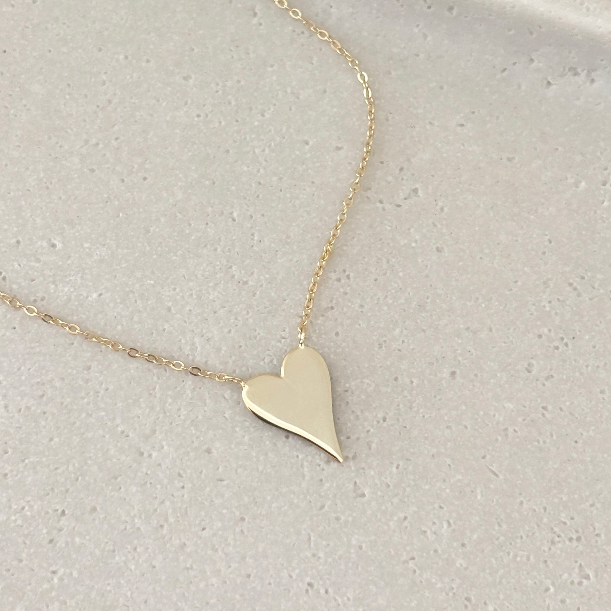 The Terry Heart Pendant Necklace, plated in 14k gold and featuring a shiny, asymmetrical heart pendant, lies on a light, textured surface.