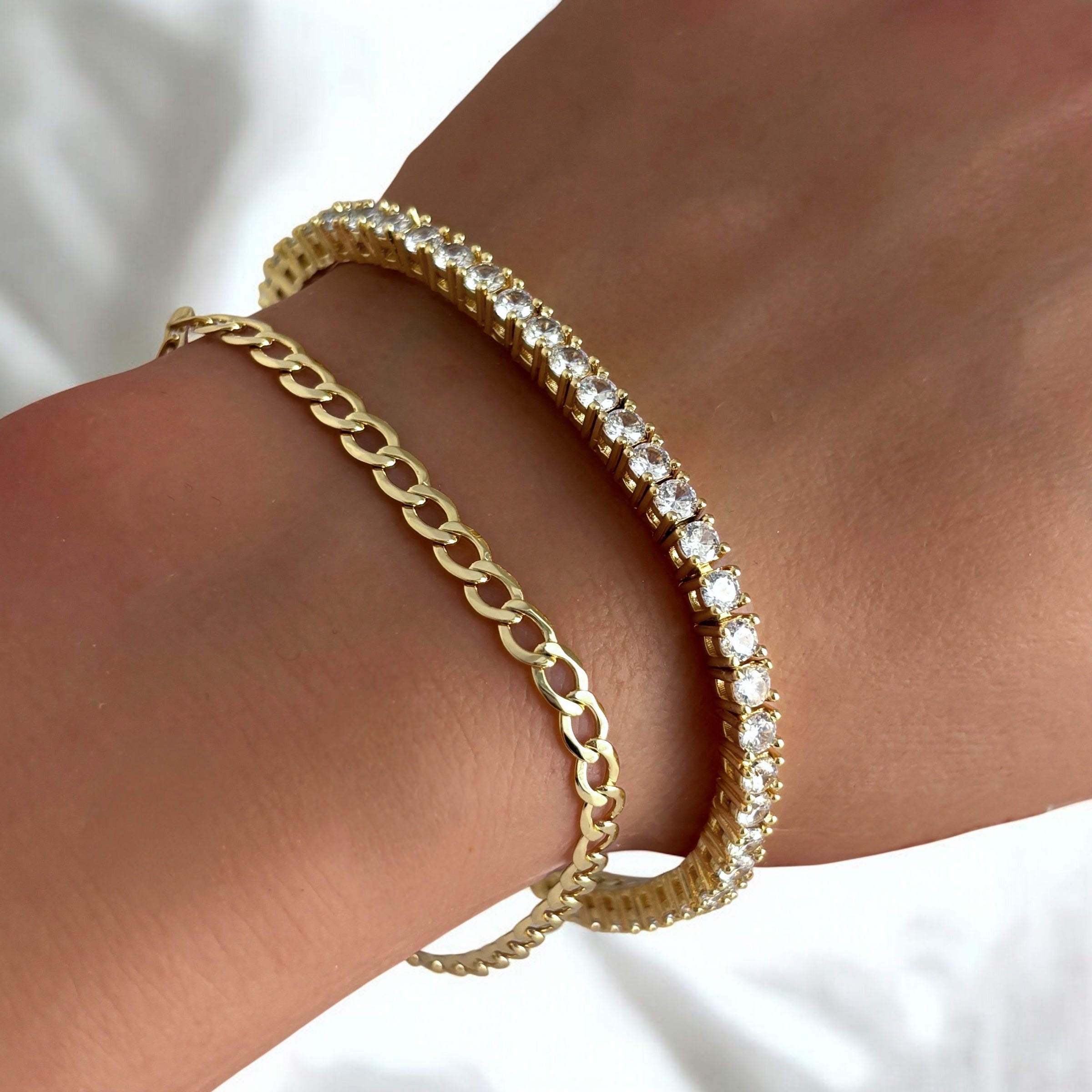 A close-up of a wrist wearing two gold bracelets: the Brynn Cuban Bracelet, a delicate 14k gold plated chain link design, paired with a dazzling tennis bracelet set with sparkling white stones.