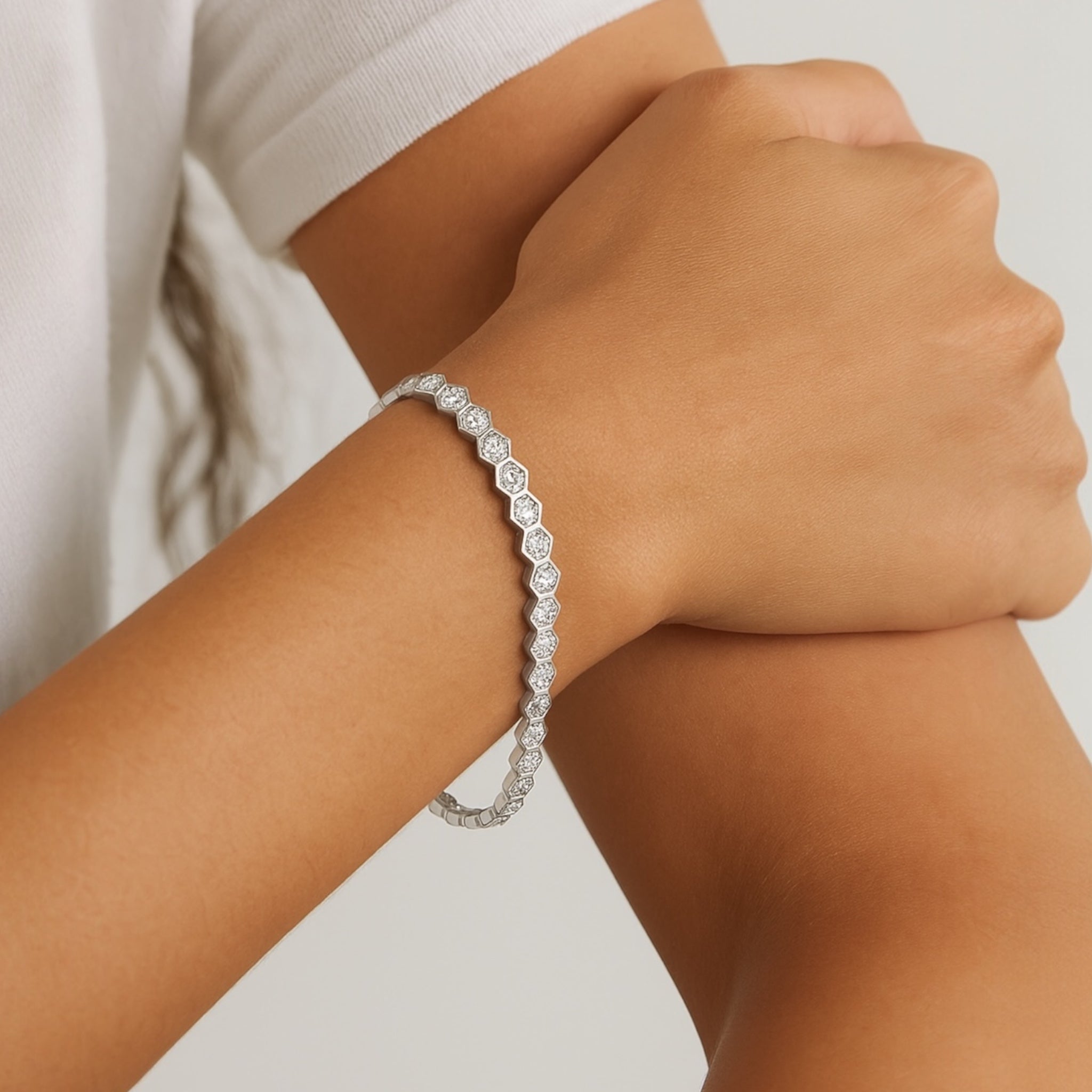 A close-up of a person’s wrist wearing the Hexagon Tennis Bracelet, featuring delicate hexagonal simulated diamond stones. Their other hand gently holds their arm. The background is neutral, and they’re dressed in a white top.