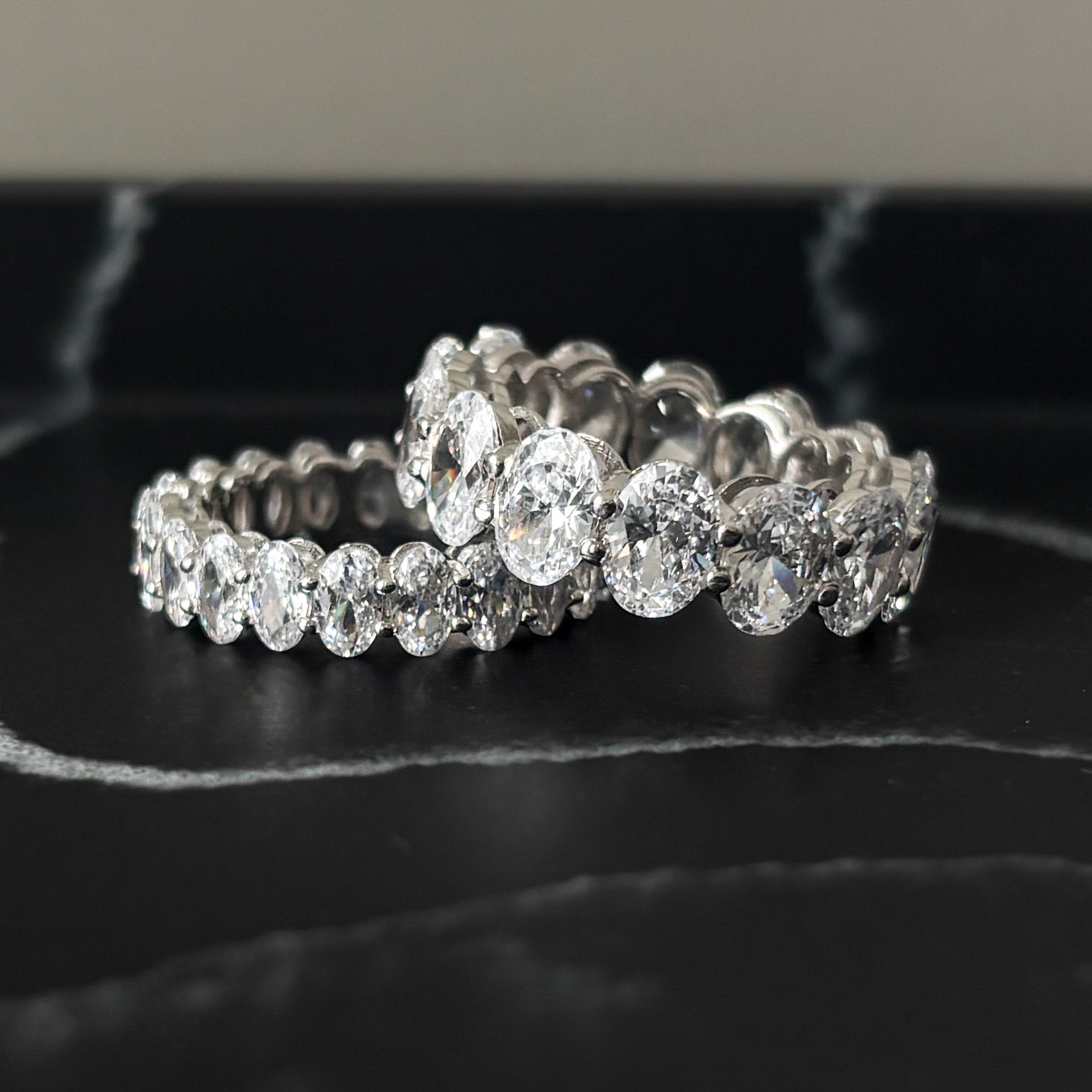 Two Bella Rings, made of 925 sterling silver with large round-cut simulated diamonds encircling each band, showcase a sparkling, luxurious look as they rest on a black marble surface.